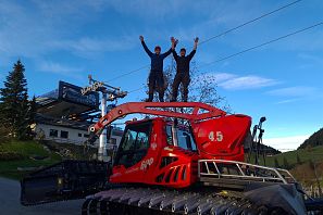 Zwei Männer stehend mit gehobenen Armen auf einer roten Pistenraupe im Sommer, im Hintergrund ist eine Liftstation und blauer Himmel
