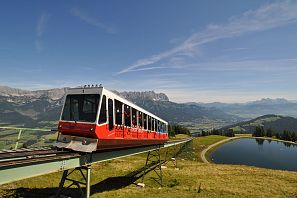 Eine rote und weiße Zahnradbahn fährt auf Schienen über eine hügelige Landschaft mit einem klaren blauen Himmel. Im Hintergrund sind Berge und ein kleiner Teich zu sehen.