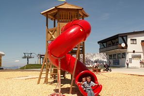 Ein Kind auf einem roten Rutschen-Element auf einem Spielplatz, während im Hintergrund ein Spielturm und ein blauer Himmel sichtbar sind.
