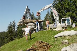Eine Baustelle in den Bergen, wo große Maschinen Steine heben. Arbeiter sind aktiv. Im Hintergrund sind Bäume und eine Berglandschaft zu sehen.