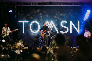 Band spielt auf einer Bühne mit großem "TOM SN" Schriftzug im Hintergrund, Publikumsmenge im Vordergrund.