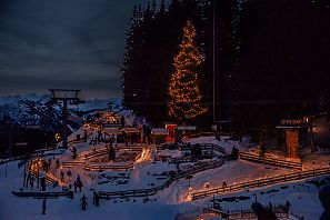 Winterlandschaft bei Nacht in den Bergen, mit einem beleuchteten Weihnachtsbaum, Schnee, Holzstrukturen und Menschen, die sich im Freien aufhalten.