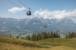 Eine Berglandschaft mit einer Seilbahn, die über grüne Wiesen und Berge führt. Im Hintergrund sind majestätische Gipfel unter blauem Himmel zu sehen.