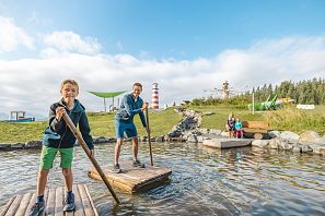Zwei Kinder stehen auf einem Floß in einem Gewässer und haben Spaß. Im Hintergrund sind Spielgeräte und ein Leuchtturm.