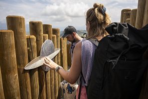 Twee mensen staan bij een houten omheining, de man kijkt en de vrouw raakt een metalen structuur aan.