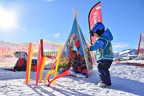 Zwei Kinder in Skikleidung spielen bei Sonnenschein vor einem bunten Tipi-Zelt im Schnee, umgeben von Skikurs-Ausrüstung und Sicherheitsnetzen