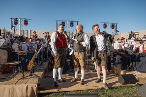 Drei Männer in traditioneller Tracht stehen auf einer Bühne, umgeben von Musikern in einer Blasmusikgruppe. Der Hintergrund zeigt eine lebhafte Atmosphäre.