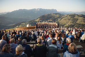 A large audience is seated on wooden benches, watching an outdoor concert on a mountain. The sun is setting behind scenic mountains in the background.