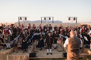 Eine Blaskapelle in traditioneller Tracht spielt draußen auf einer Bühne. Musiker stehen in Reihen, im Hintergrund sieht man Berge und eine abendliche Landschaft.