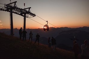 Una góndola de teleférico ascendiendo al atardecer, con siluetas de personas en la ladera de la montaña. El paisaje muestra montañas en el horizonte, iluminadas por el sol.