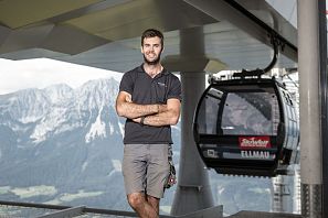 Ein lächelnder Mann in einem schwarzen T-Shirt steht mit verschränkten Armen vor einer Seilbahn. Im Hintergrund sind hohe Berge und ein klarer Himmel zu sehen.