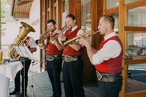 Die Bläsergruppe der Musikkapelle Westendorf in roten Westen spielt Musik auf einer Terrasse mit Holzfassade im Hintergrund.