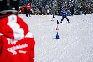Ein Kind mit blauer Jacke fährt Ski durch blau-orange Kegel. Ein Skilehrer in Rot schaut zu. Schneebedeckte Bäume im Hintergrund. Klarer Wintertag.
