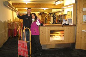 Zwei Personen stehen in einem Holzraum mit einem Empfangstresen. Die Frau trägt eine lila Jacke, der Mann hebt einen Schlitten. Um sie herum sind verschiedene Ausrüstungsgegenstände.