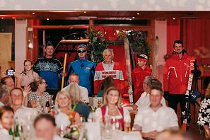 Männer in nostalgischer und moderner Skibekleidung vor einem roten, dekorierten Lift in einem Festsaal.