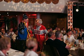Zwei Männer in nostalgischer Skibekleidung unterhalten sich vor einem sitzenden Publikum.