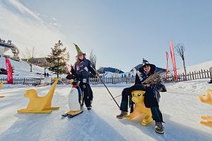 Zwei Personen stehen auf einer Eisfläche. Eine Person trägt einen Hut und hält eine Skiausrüstung, während die andere mit einem Tier-Motiv auf einer gelben Schlittenfigur sitzt. Hintergrund: schneebedeckte Landschaft.