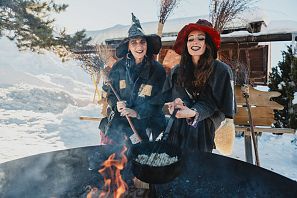 Zwei Frauen in ungewöhnlichen Kostümen stehen vor einem großen Feuer in einer verschneiten Landschaft. Eine kocht in einem Kessel, während die andere lächelt und sie anfeuert.