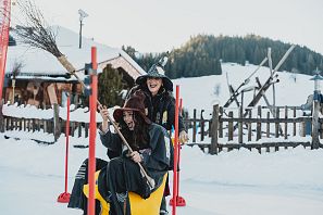 Zwei Frauen in Hexenkostümen sitzen fröhlich auf einem gelben Schlitten im Schnee. Sie lachen und haben Besen in der Hand, während eine winterliche Landschaft im Hintergrund zu sehen ist.