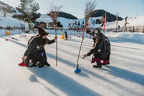 Zwei Personen in winterlicher Kleidung spielen auf einer zugefrorenen Fläche. Sie tragen Hüte und nutzen kleine Besen, um bunte Scheiben zu bewegen. Winterlandschaft im Hintergrund.