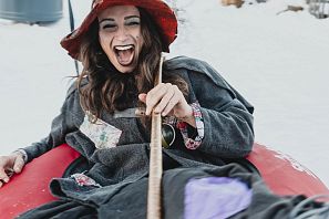 Eine fröhliche Frau mit einem roten Hut sitzt auf einem roten Gummireifen im Schnee. Sie lacht laut und hält einen Stock in der Hand. Im Hintergrund sind verschneite Bäume zu sehen.
