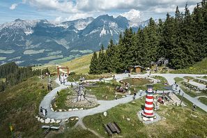 Ein panoramischer Blick auf eine Berglandschaft mit Spielmöglichkeiten im Vordergrund. Menschen genießen die Natur, umgeben von Bäumen und Bergen.