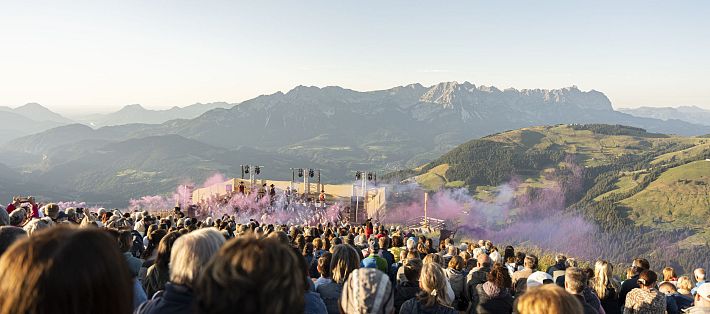 A large crowd gathered at a mountaintop concert, enjoying the performance with a scenic backdrop of mountains and colorful smoke rising into the sky.