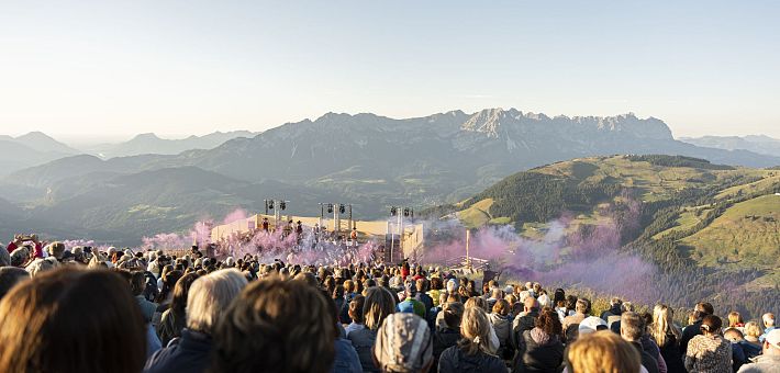 Una gran multitud de personas mira un espectáculo en la cima de una montaña con vistas a un paisaje montañoso y niebla de colores en el aire.