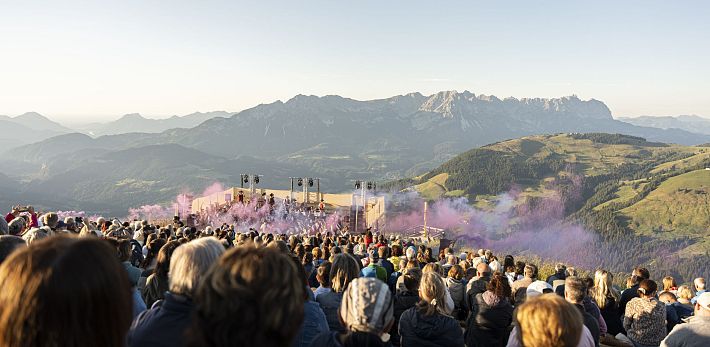 Eine Menschenmenge auf einem Berg schaut auf eine Bühne, mit Bergen im Hintergrund und lilanem Rauch in der Luft.