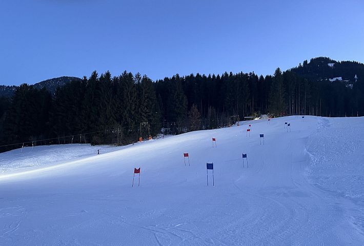 En skibakke med flag til at markere ruter og omgivende skove, badet i blødt morgenlys, der skaber en fredelig vinterstemning.