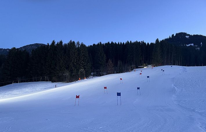 A snowy ski slope at dusk, marked with red and blue flags. The slope is bordered by dense pine trees and under a clear sky.