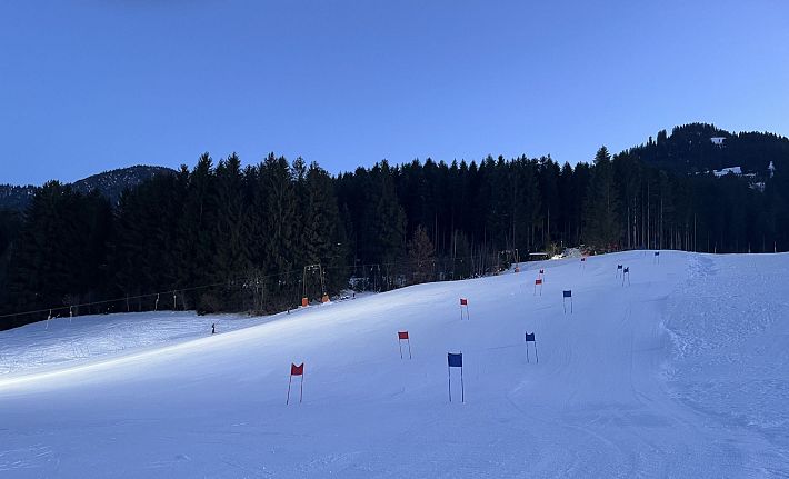 Pista de esquí al amanecer con nieve fresca, banderas de colores y un bosque en el fondo, bajo un cielo claro y azul.