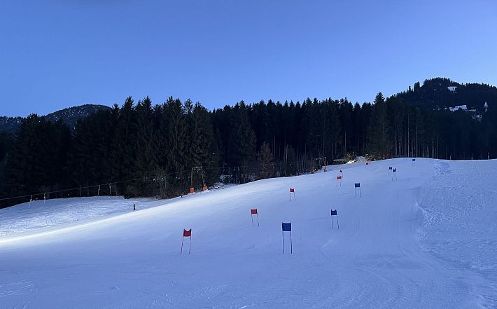Zasněžená sjezdovka osazená slalomovými brankami modré a červené barvy. Kolem jsou lesy, nad nimiž se klene stmívající se obloha.