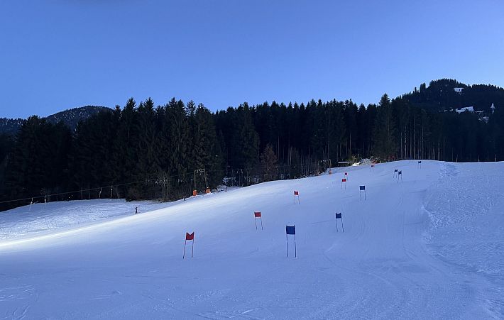 Sneeuwhelling omgeven door een dicht bos bij schemering, met blauwe en rode vlaggen langs het parcours, onder een heldere hemel.