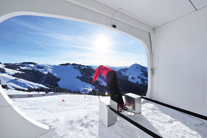 Person auf Skisprungschanze vor schneebedeckten Alpen unter blauem Himmel. Die Sonne scheint hell, und das Bild hat einen futuristischen Rahmen.
