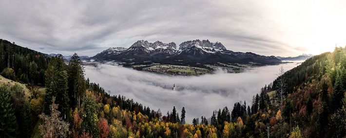 Eine atemberaubende Landschaft mit majestätischen Bergen im Hintergrund. Dichte Nebelschwaden durchziehen das Tal, während sich bunt gefärbte Bäume im Vordergrund zeigen.