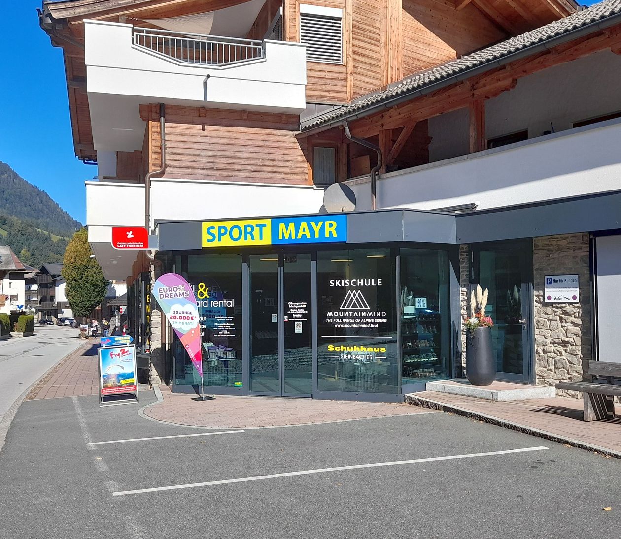 The image shows a modern storefront with large glass windows and a sign that reads "SPORT MAYR." The building has a wooden facade and is located in a scenic area with mountains visible in the background.
