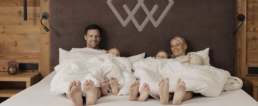 Mutter und Vater mit zwei Kindern liegen zugedeckt im Doppelbett im Hotel Kaiserlodge während Füße und Gesichter zu sehen sind