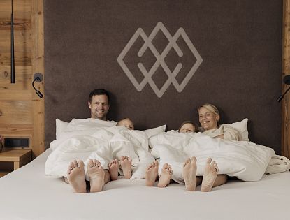 Mutter und Vater mit zwei Kindern liegen zugedeckt im Doppelbett im Hotel Kaiserlodge während Füße und Gesichter zu sehen sind