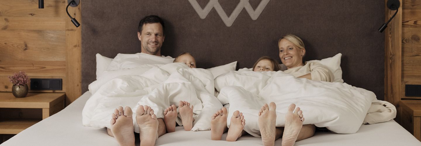 Mutter und Vater mit zwei Kindern liegen zugedeckt im Doppelbett im Hotel Kaiserlodge während Füße und Gesichter zu sehen sind