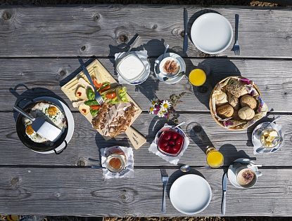 Eine rustikale Holztischplatte mit einer Brotzeitplatte, frischem Gemüse, einem Getränk, Dip und Brot. Blumen und Tageslicht vervollständigen die Szene.