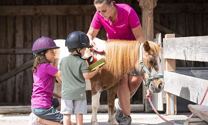 Zwei Kinder mit Reithelmen stehen neben einem kleinen braun-weißen Pony, begleitet von einer Betreuerin in pinkem Shirt, vor einem Holzstall.