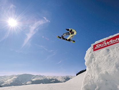 Snowboarder springt im Funpark Boarders Playground vor blauem Himmel und verschneiten Alpen.