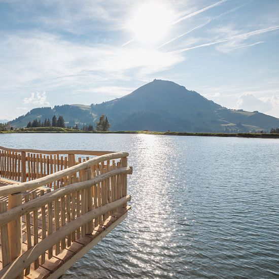 Holzsteg am Brantlalmsee in der RiesenWelt SkiWelt Wilder Kaiser Brixental, Blick über den See auf eine Berglandschaft bei Sonnenschein
