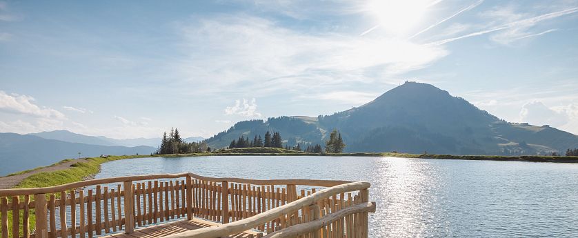 Holzsteg am Brantlalmsee in der RiesenWelt SkiWelt Wilder Kaiser Brixental, Blick über den See auf eine Berglandschaft bei Sonnenschein