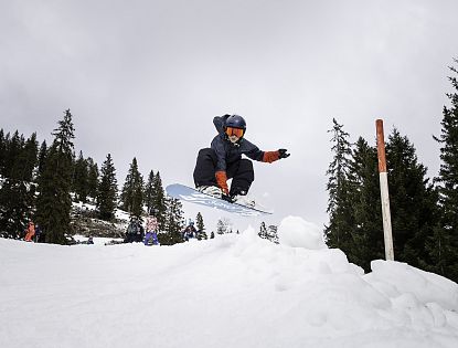 Snowboarder springt in der Luft über eine Schneerampe im Funpark, umgeben von anderen Wintersportlern und verschneiten Tannen