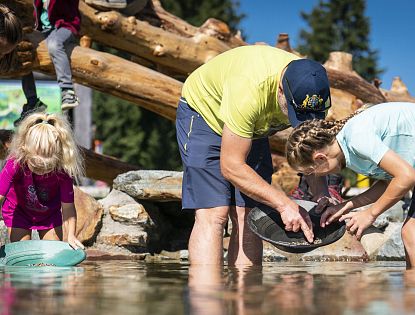 Ein Mann und zwei Kinder stehen im flachen Wasser in der Ellmis Zauberwelt und sieben mit runden Schalen Steine oder Sand. Im Hintergrund sind Holzstämme und Bäume zu sehen.