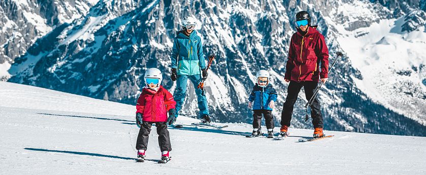 Eine Familie steht auf einer schneebedeckten Piste in den Bergen. Zwei Kinder in bunten Ski-Anzügen und Helmen schauen lächelnd, während die Eltern bereit sind zum Skifahren.