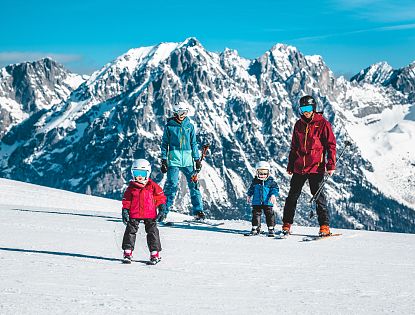 Eine Familie steht auf einer schneebedeckten Piste in den Bergen. Zwei Kinder in bunten Ski-Anzügen und Helmen schauen lächelnd, während die Eltern bereit sind zum Skifahren.