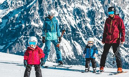 Familie in Skiausrüstung auf einer verschneiten Piste, umgeben von beeindruckenden Berggipfeln. Kinder tragen bunte Jacken, alle tragen Helme.